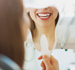 Mulher sentada na cadeira do dentista, sorrindo e segurando um espelho com o reflexo do seu sorriso